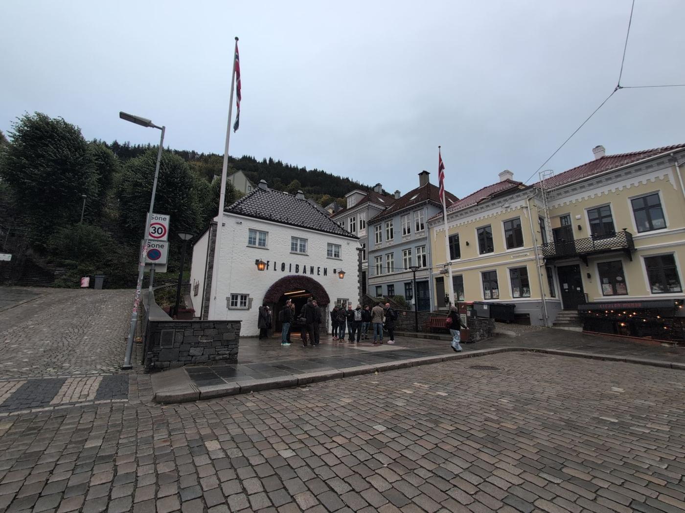 Cobbled lane rising to the white Fløibanen funicular station house, flags on poles, a few people by the entrance.