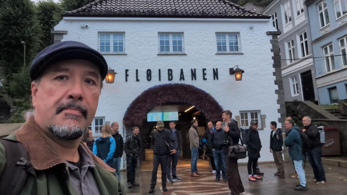 Selfie of a man in a flat cap with a grey goatee, the Fløibanen station entrance and the waiting group behind him.