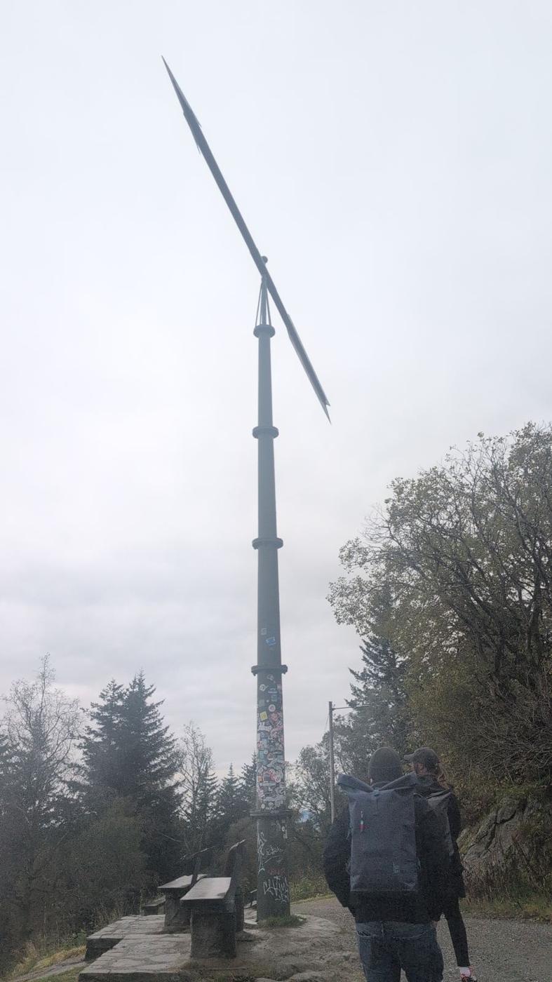 A tall slender mast topped by a long single blade rises into a white sky above spruce trees, a walker with a backpack in the foreground.