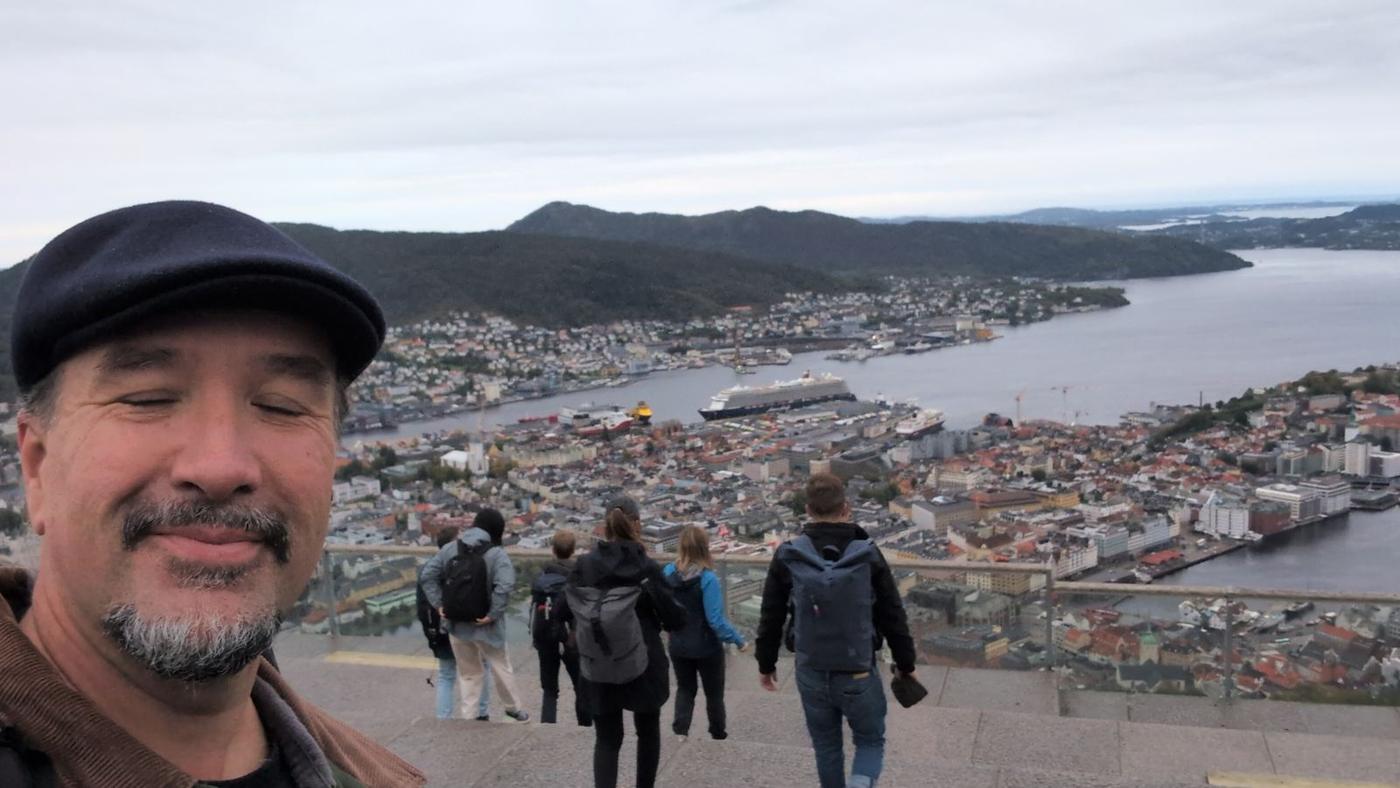 Selfie of the same man in a flat cap with Bergen's harbour, a docked cruise ship and the fjord below, walkers on the viewing terrace.