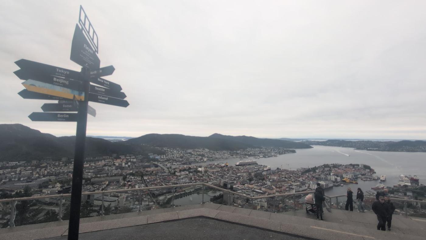 Wide view from Fløyen over Bergen, the harbour and the fjord to distant hills, with the signpost at left.